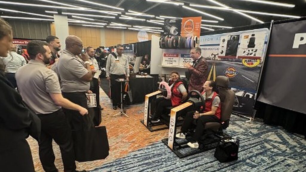 Joe Turner performing magic for two guests seated in driving simulators at a trade show booth while a crowd watches.